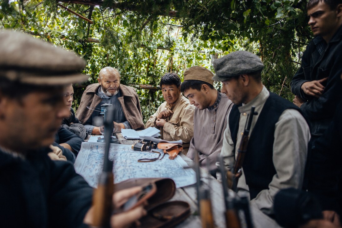 Group of men gathered around a table outdoors examining documents under hanging vegetation.