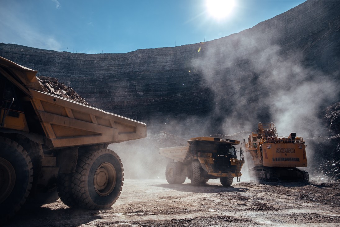 Large yellow Belaz trucks and excavator extracting ore in an open pit mine with dust clouds.