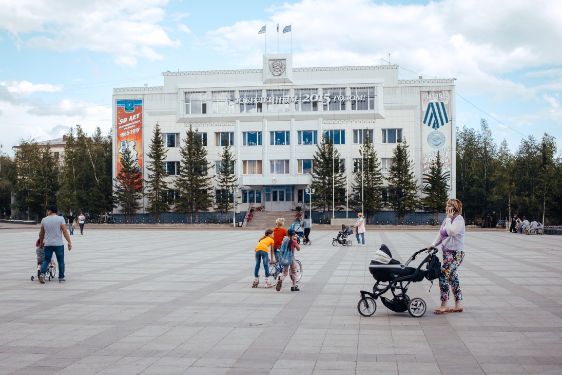 A white Soviet-style government building with blue trim overlooking a large plaza with children playing and adults walking.