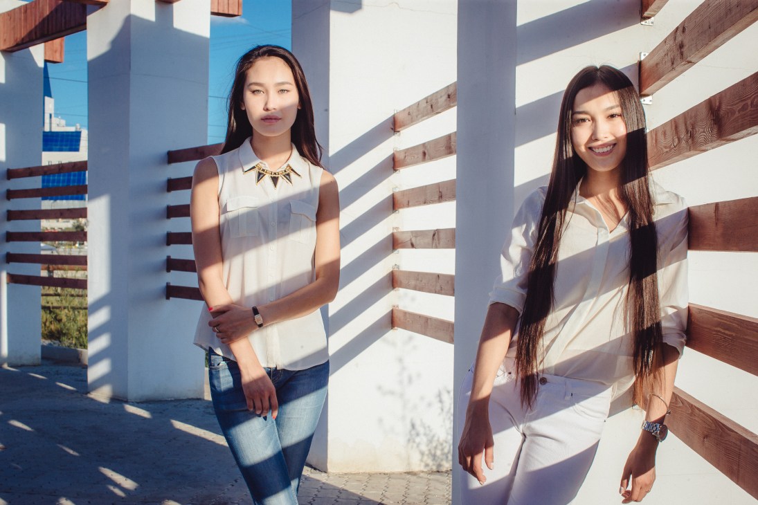 Two young women stand against a wooden fence structure in bright sunlight, wearing light-colored tops.