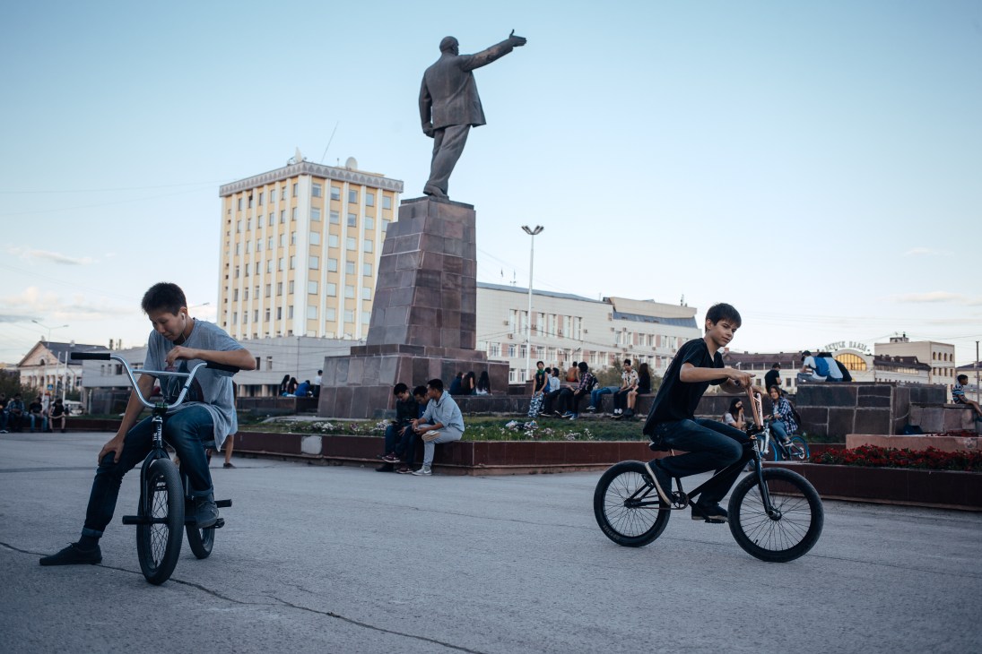 Teenagers riding bikes in Lenin Square, Yakutsk's central square with statue and Soviet-era buildings.