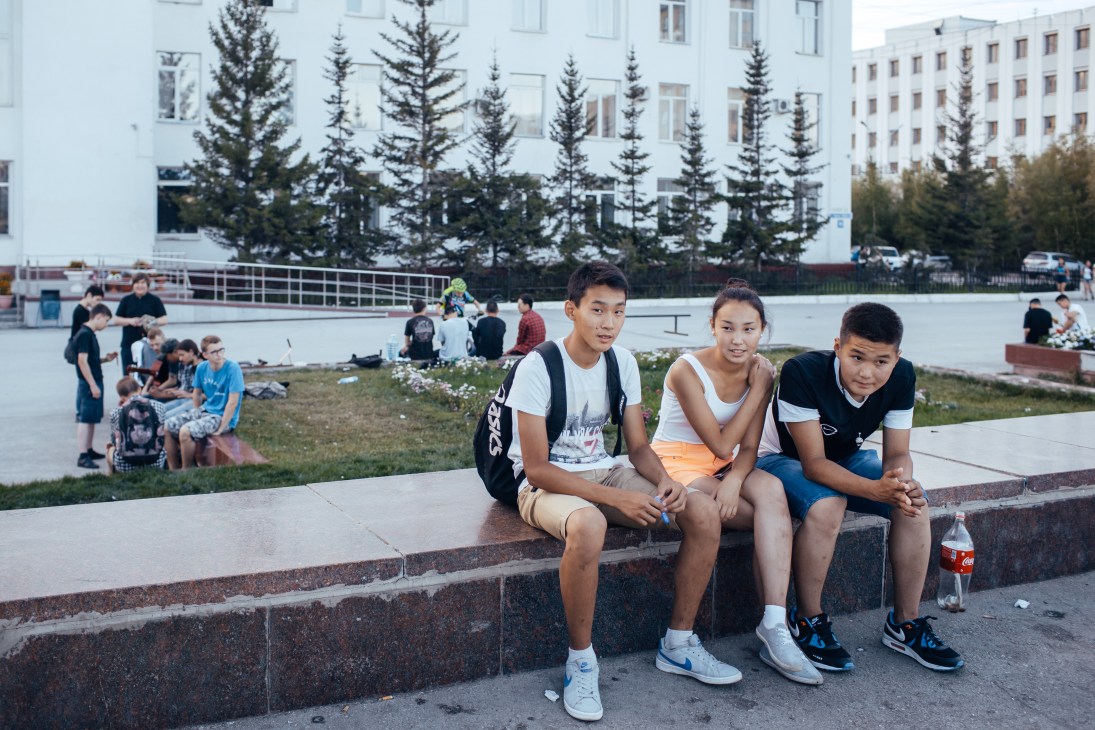 Three teenagers sit on a concrete bench in a public square with Soviet-era apartment buildings and evergreen trees in the background.