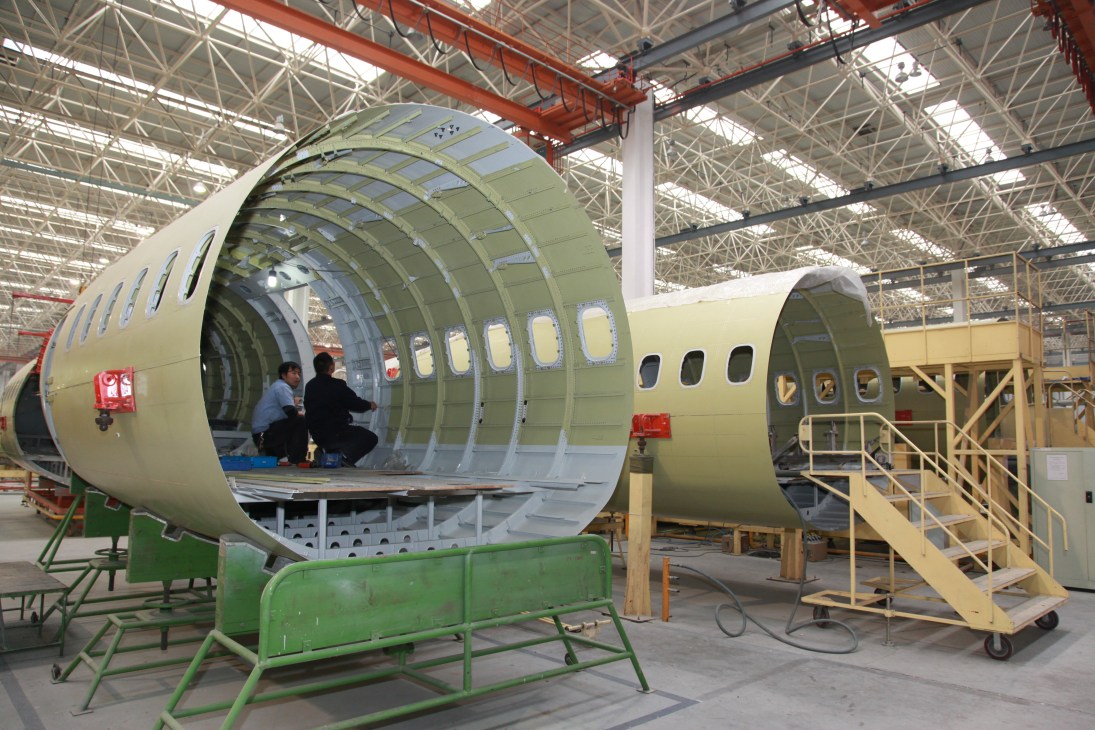 Workers inside a large aircraft fuselage section in an industrial factory setting.