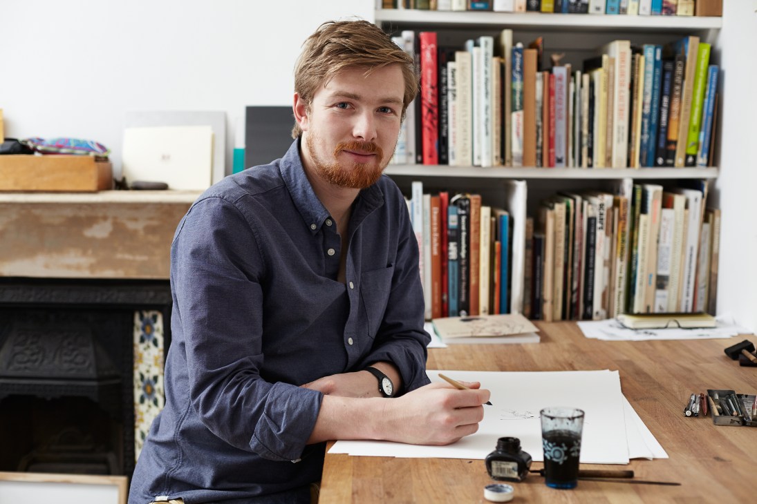 Artist George Butler sits at a wooden desk with drawings and ink, bookshelves behind him.