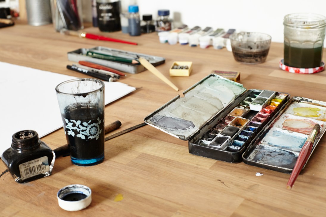 Artist's workspace with ink bottles, watercolor palette, brushes, pencils, and water cups on a wooden desk.