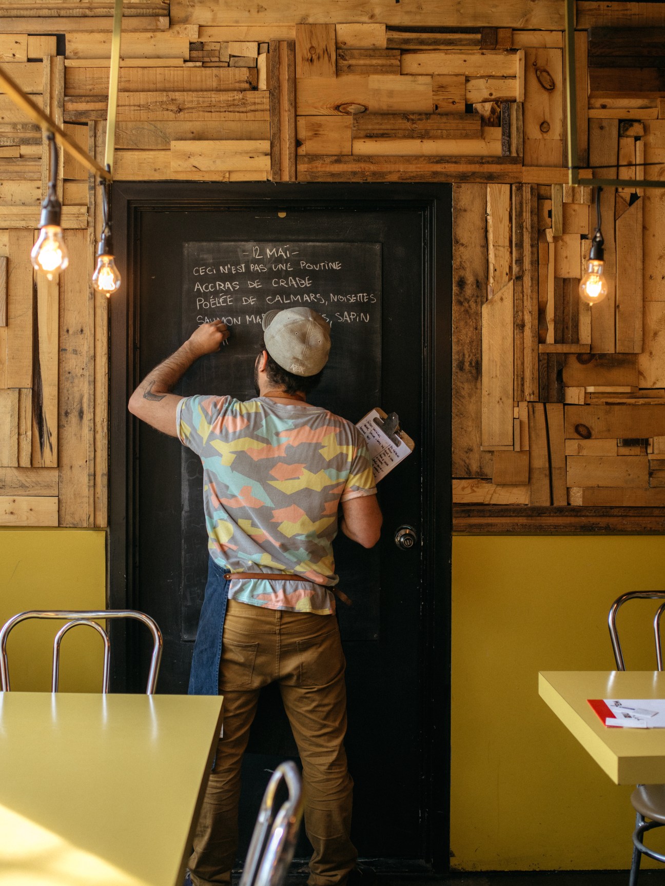A man in a colorful shirt and white hat writing daily specials on a blackboard menu in a restaurant.