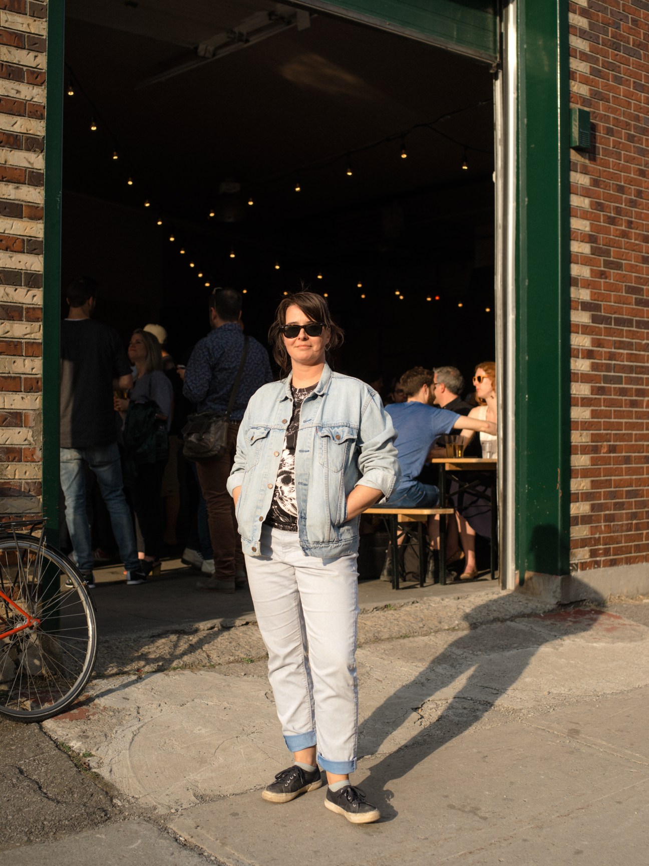 Bernadette Houde standing outside Alexandraplatz bar entrance at dusk wearing denim jacket and sunglasses.