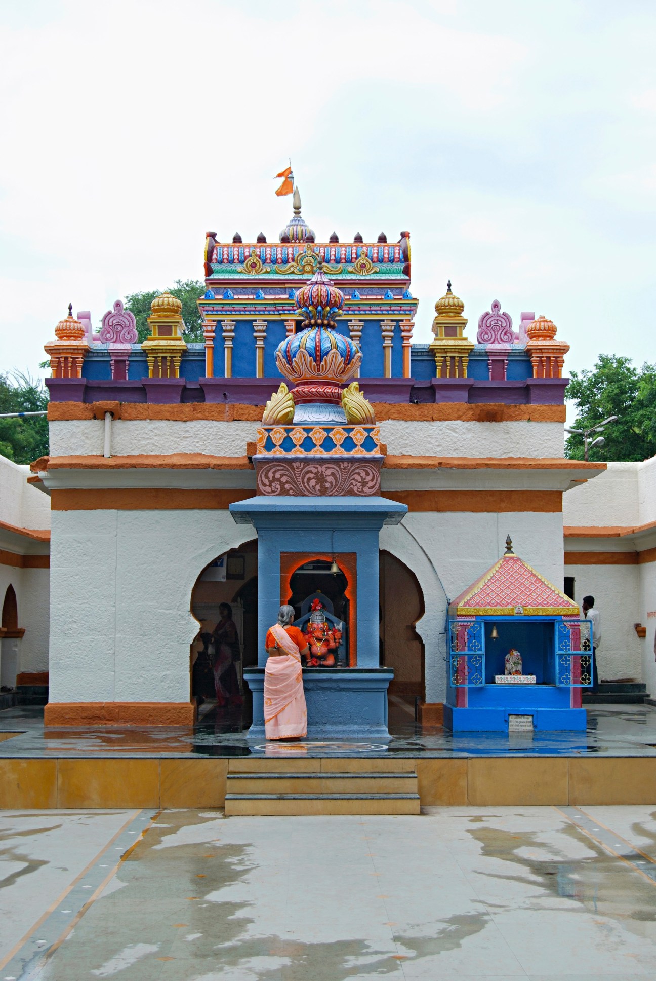 Colorful Hindu temple with blue and orange ornamental architecture and a woman in pink sari standing in front.