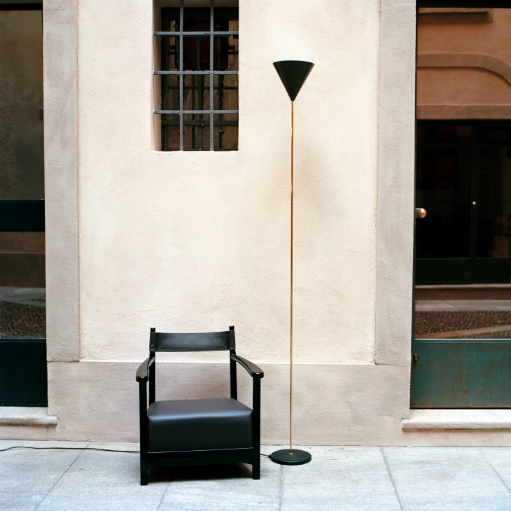 Black Superslim Imbuto floor lamp with brass stem next to a wooden Chinotto armchair against a cream-colored wall.