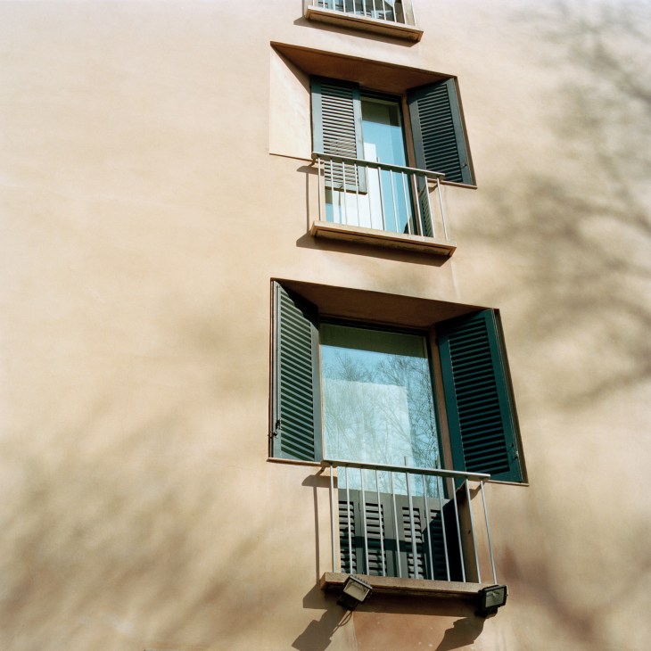 Recessed windows with angled green shutters and metal balconies on a cream-colored building facade.