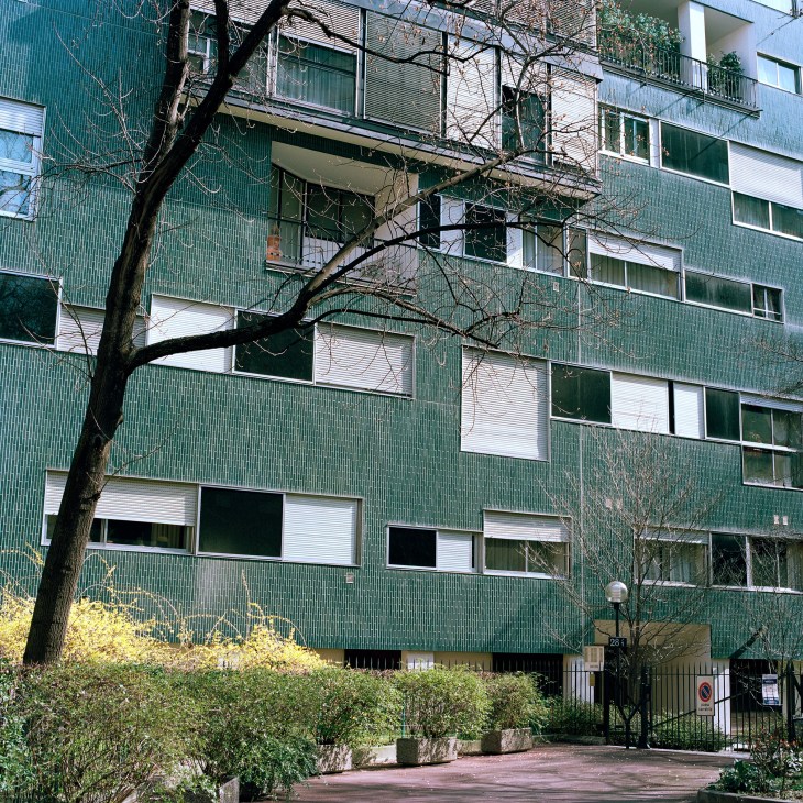 Green clinker tile apartment building with varying window sizes and a bare tree in front, Milan