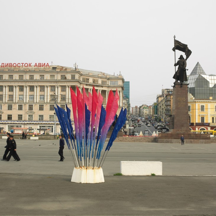 Colorful decorative sculpture with blue and red ribbons on a square in Vladivostok, with historic buildings and a statue in the background.