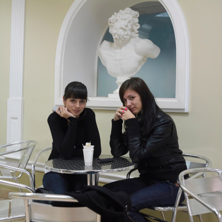 Two students sit at a modern table in a university café with a classical bust sculpture displayed in an arched alcove behind them.
