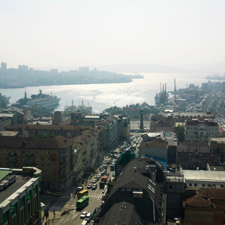 Aerial view of Vladivostok showing the harbour with military and commercial ships, Soviet-era buildings, and the Golden Horn inlet.