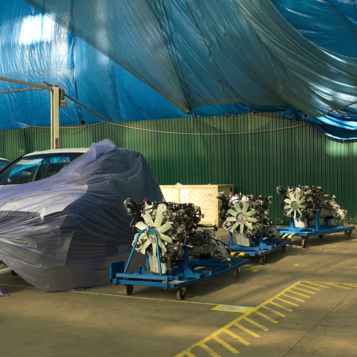 Car engines on blue trolleys at Sollers factory in Vladivostok, with a covered vehicle and green corrugated metal walls.