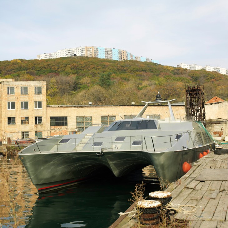 Modern luxury catamaran docked at old military shipyard in Vladivostok with Soviet-era apartment buildings on hillside.