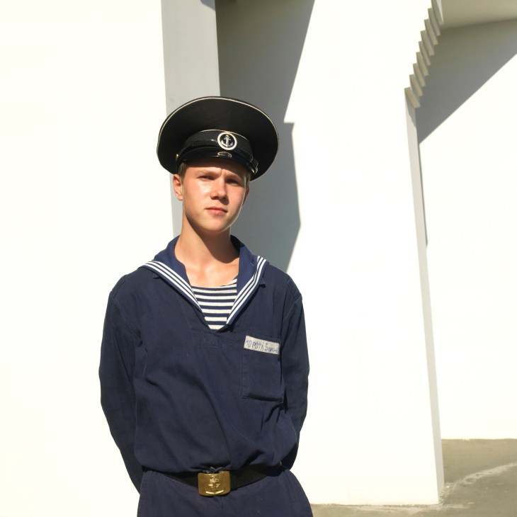 Russian naval serviceman in uniform standing against concrete structure.