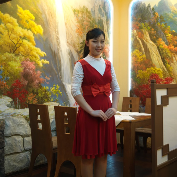 Young woman in red dress with bow standing in North Korean restaurant with painted landscape mural backdrop.