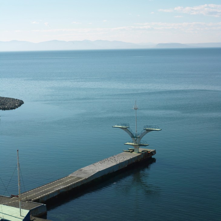Soviet-era diving platform extending into calm blue water with mountains visible across the bay.