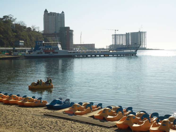 Central beach in Vladivostok with orange pedal boats on sand, ferry boat and high-rise buildings under construction in background.