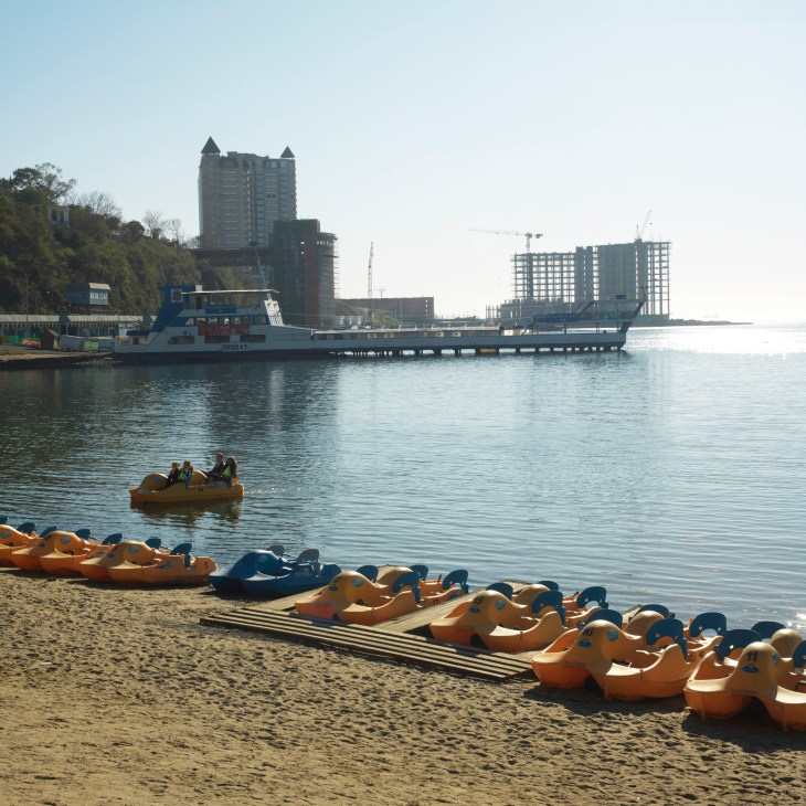 Central beach in Vladivostok with orange pedal boats on sand, ferry boat and high-rise buildings under construction in background.