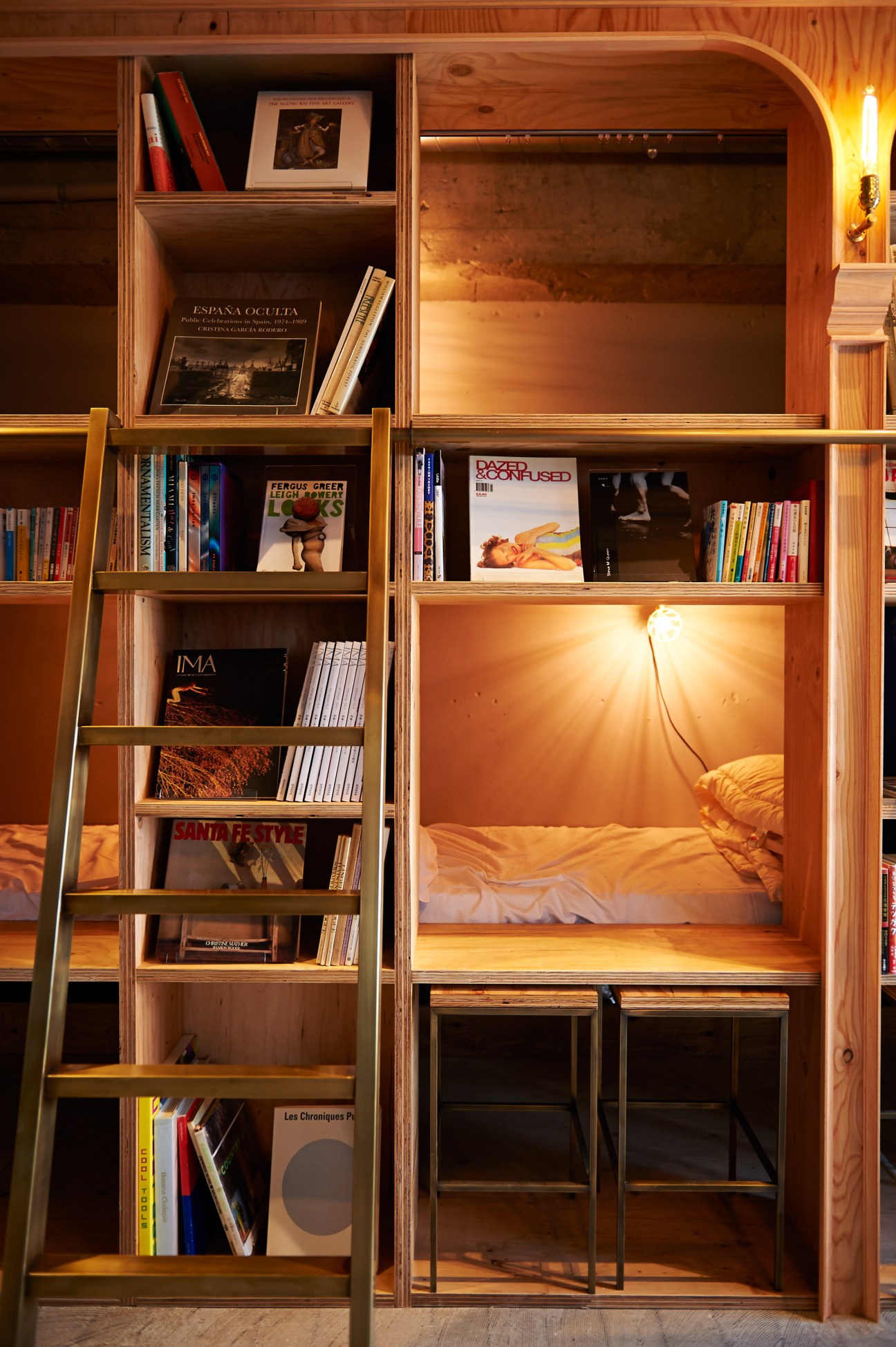 A wooden sleeping pod built into bookshelves with books lining the compartments and warm lighting inside.