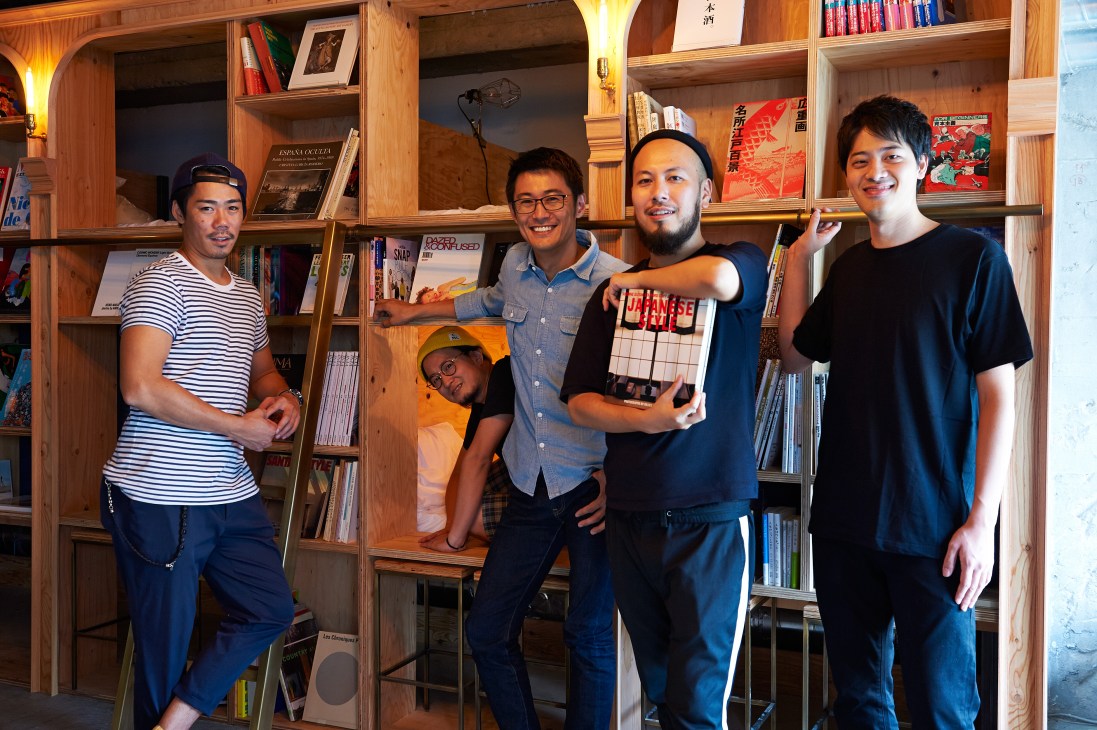 Five people stand smiling in front of wooden bookshelves filled with books and magazines at Book and Bed Tokyo hostel.