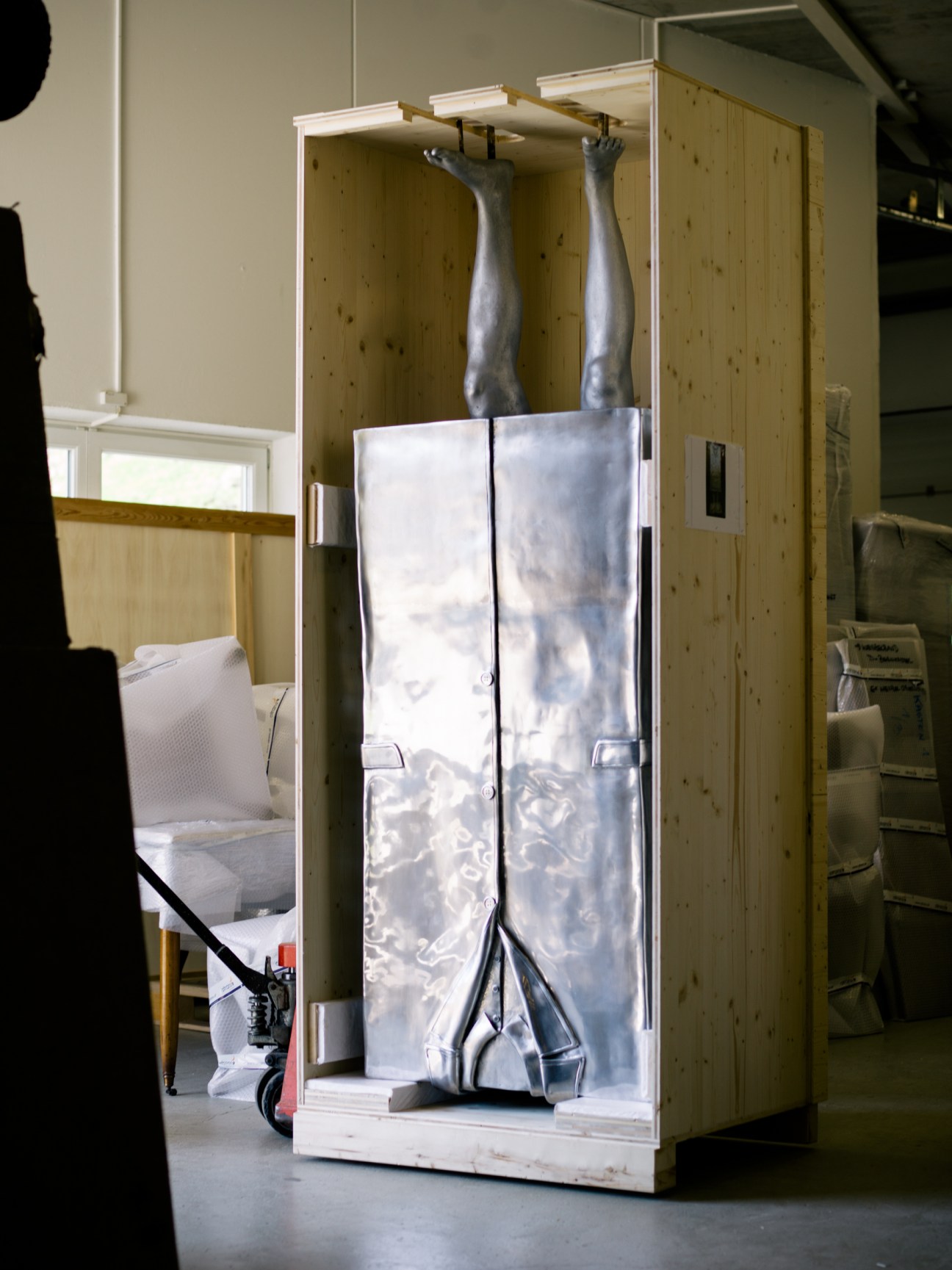 Erwin Wurm sculpture with raised legs visible inside a wooden crate with white padding and translucent plastic sheeting.