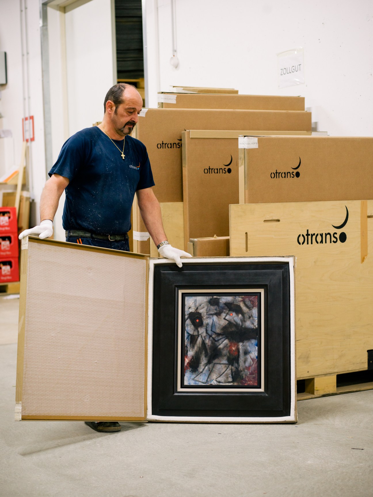 Worker in Otrans warehouse handling a framed painting wrapped in protective materials, surrounded by cardboard shipping crates.