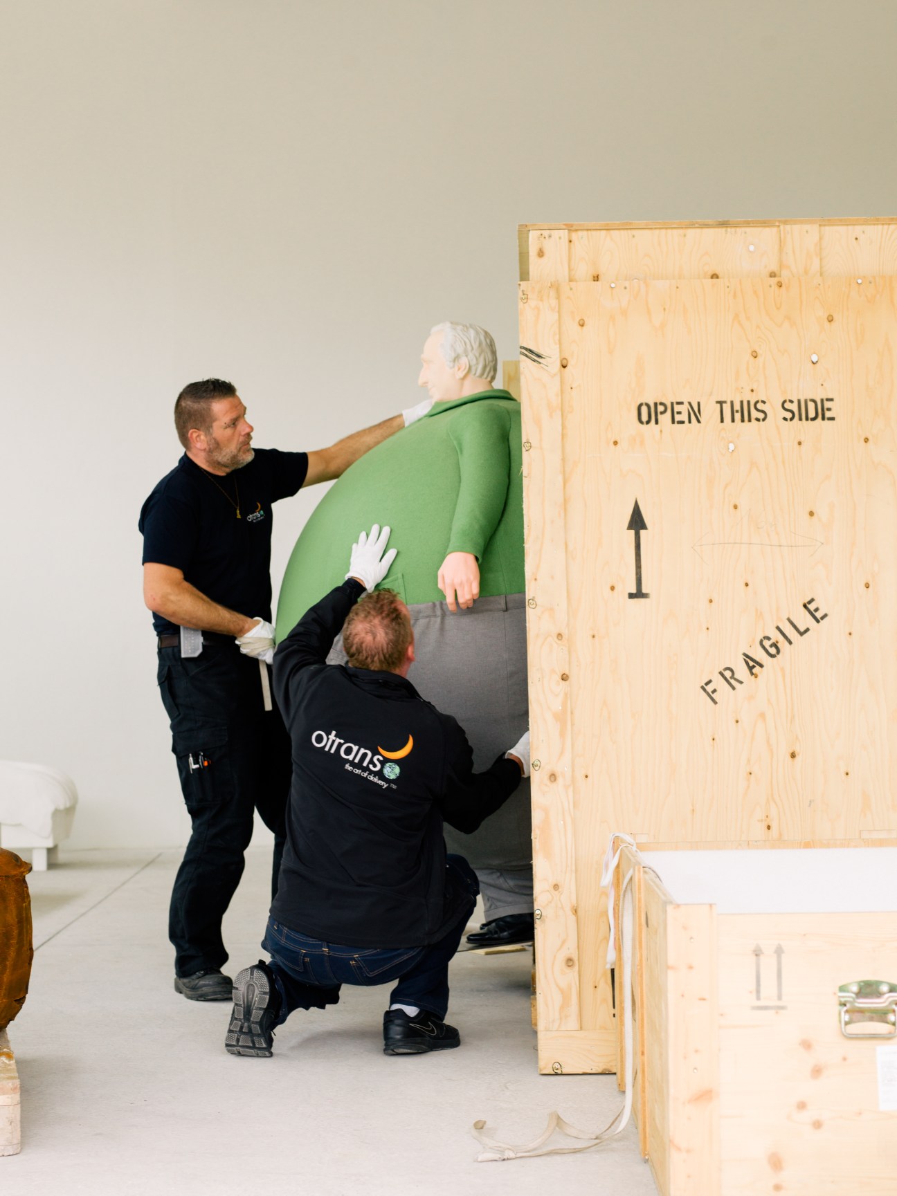Two Otrans workers carefully placing an Erwin Wurm sculpture into a wooden shipping crate marked 'OPEN THIS SIDE' and 'FRAGILE'.