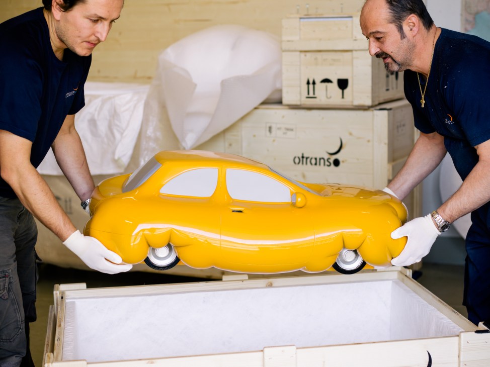 Two workers in navy Otrans uniforms carefully handle a bright yellow car-shaped sculpture by Erwin Wurm into a foam-lined wooden crate.
