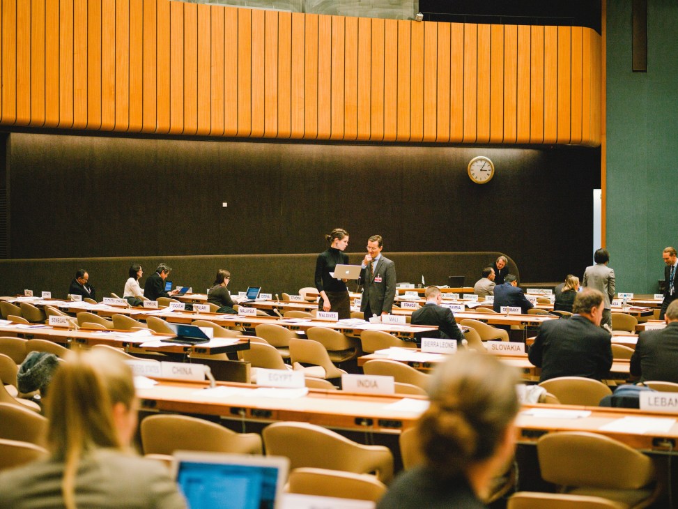UN delegates discuss landmine treaty in a large conference room with country name placards at wooden desks.
