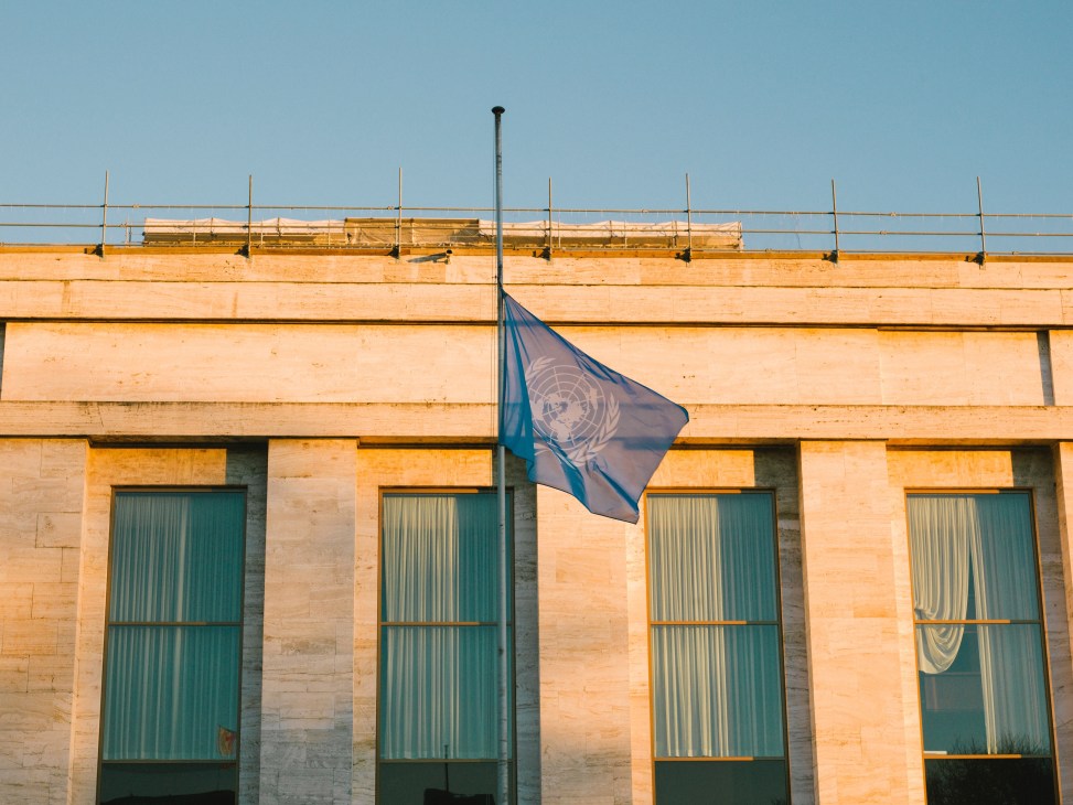 UN flag flying outside the Palais des Nations building in Geneva with sandstone facade and green doors.