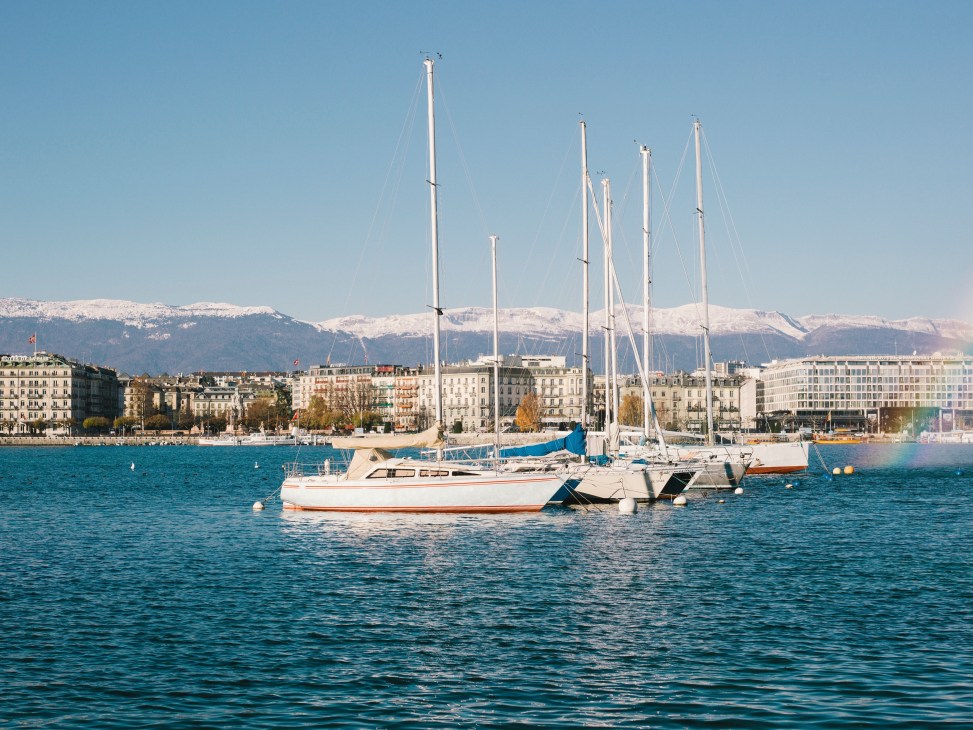 Sailboats moored on Lake Geneva with snow-capped Alps and lakeside city buildings in background.