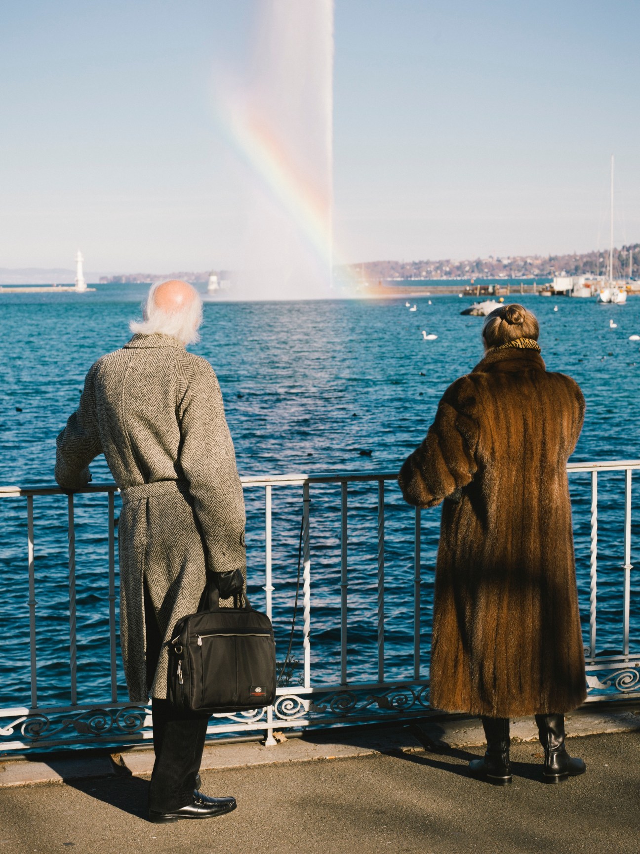 Two residents viewing Lake Geneva and the Jet d'Eau fountain from a lakeside promenade.