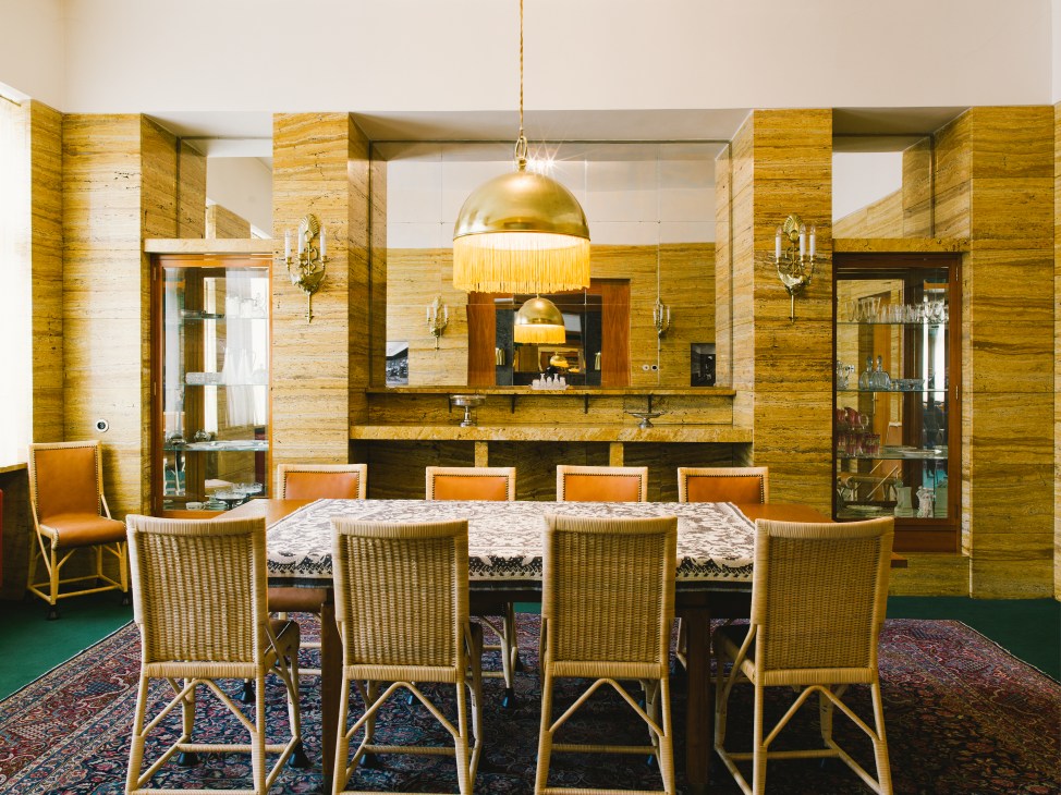 Restored dining room with travertine walls, brass pendant light, woven chairs and built-in cabinetry at Klatovská 12.