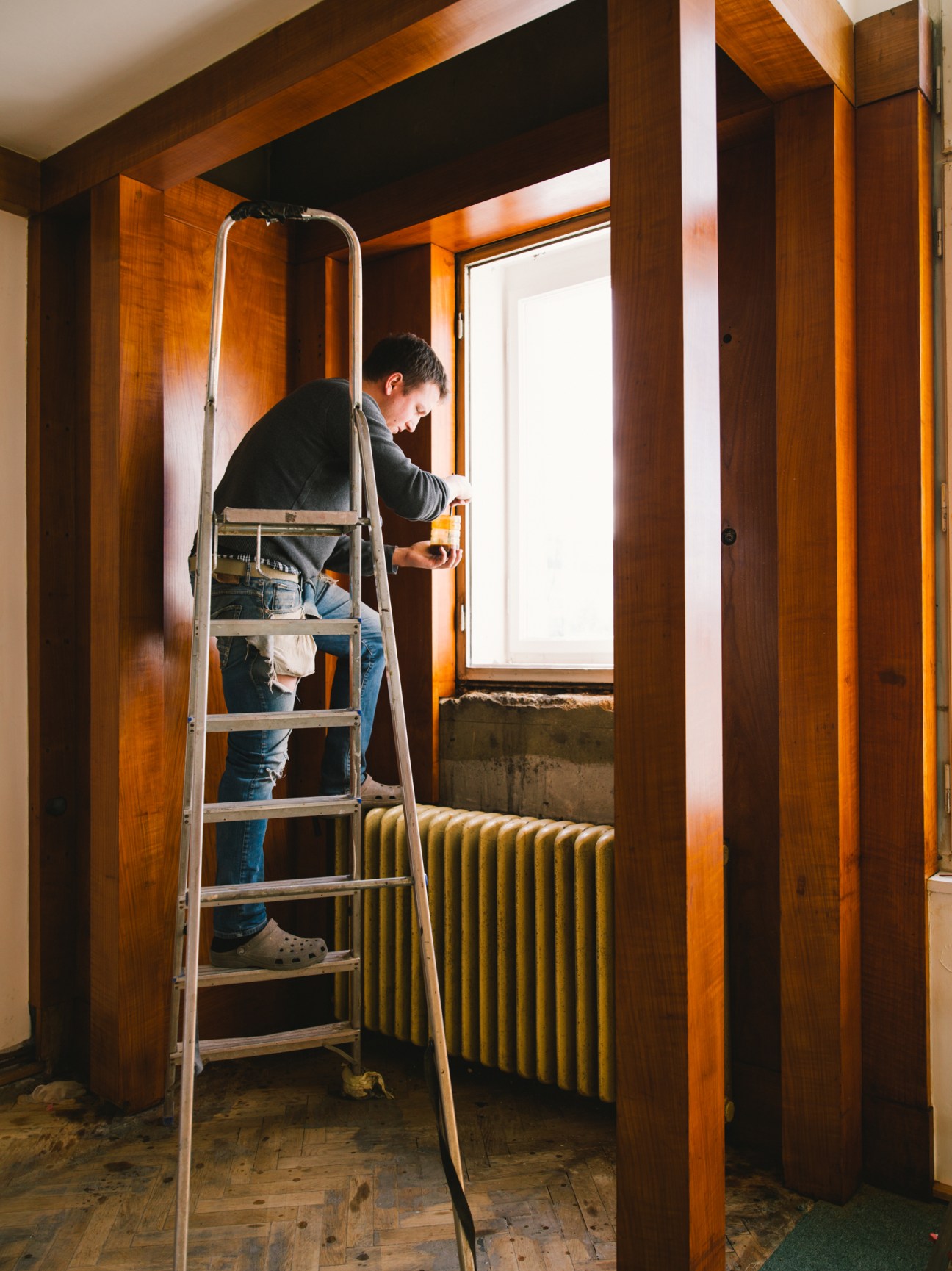 Worker on ladder varnishing mahogany wood panelling in a bedroom with a yellow radiator visible.