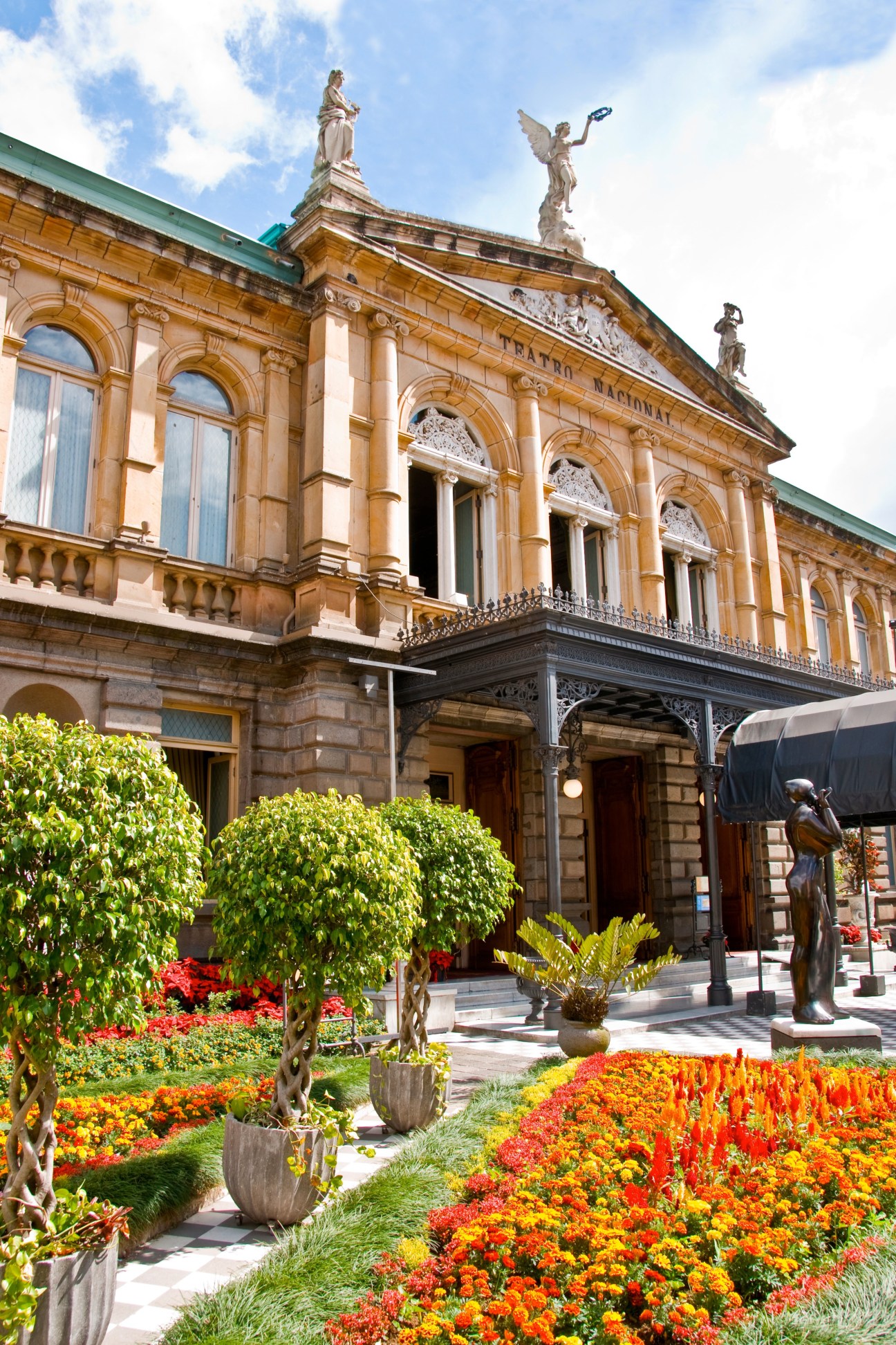 San José's National Theatre with neoclassical architecture, golden stone façade, statues, and colorful flower gardens.