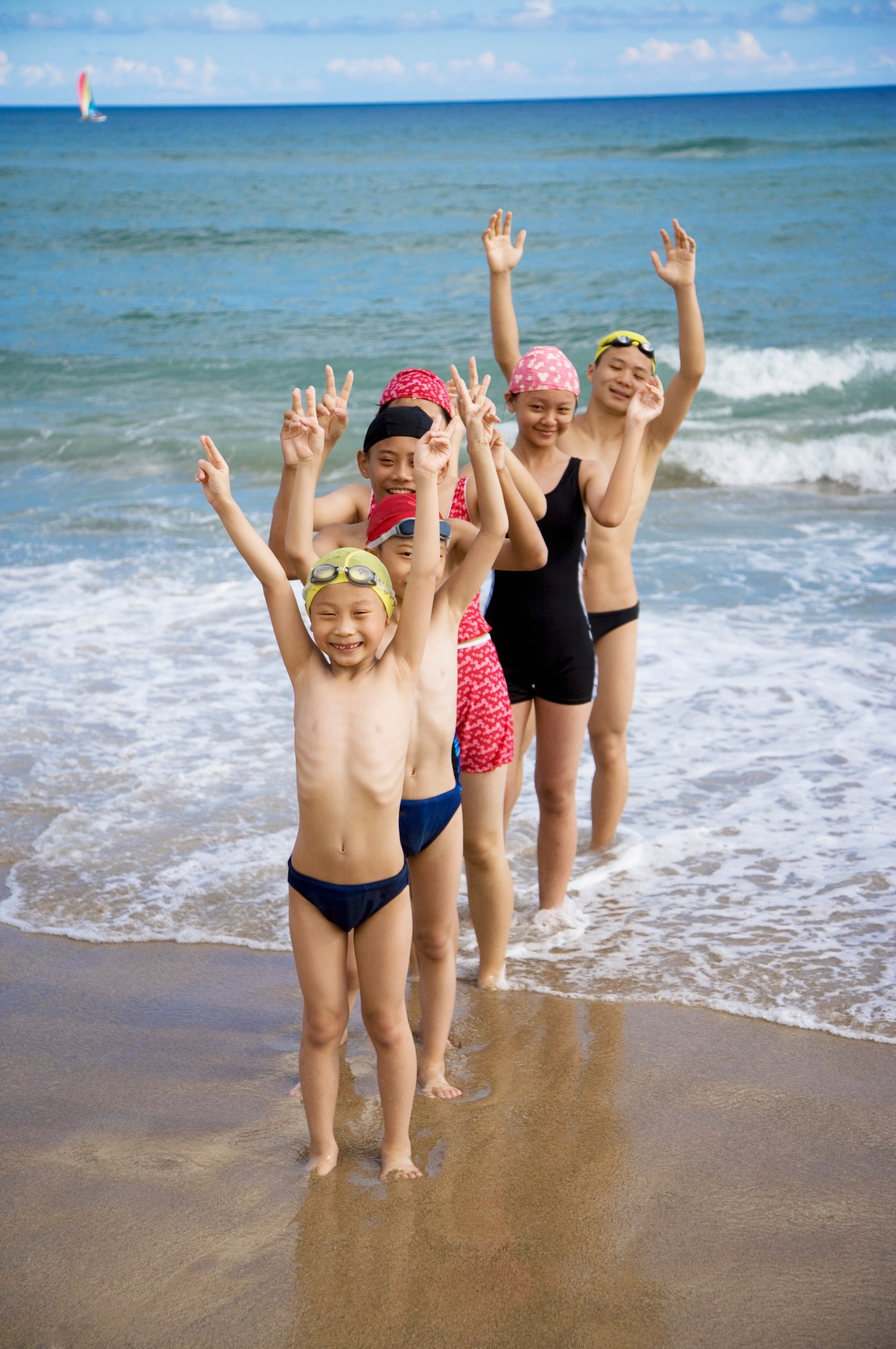 Children in swimming costumes and caps standing in the ocean waves with their arms raised.