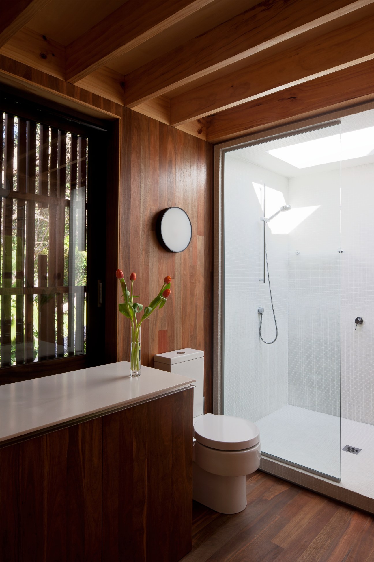 Modern ensuite bathroom with wood-paneled walls, white toilet, glass shower enclosure, and round mirror.