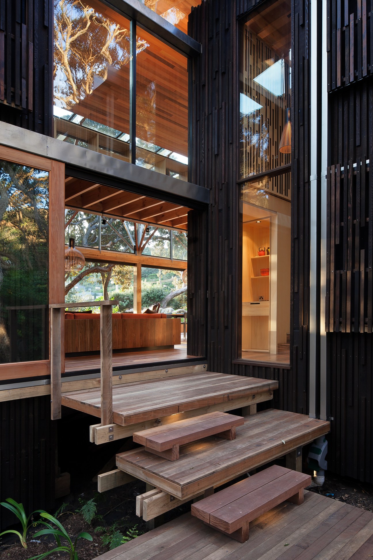 Wooden steps leading to a breakfast deck on a modern beach house with black-stained wood cladding and large glass windows.