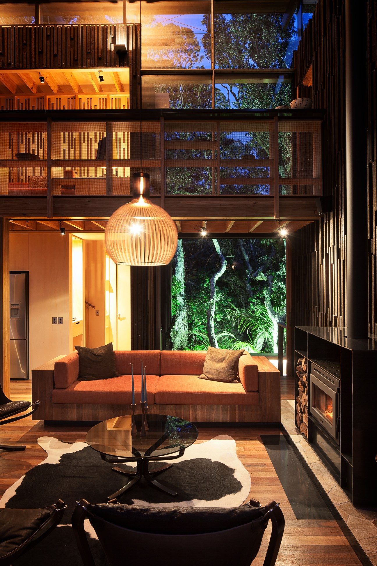 Modern wood-lined living room with orange sofa, pendant lamp, and large windows overlooking forest at night.