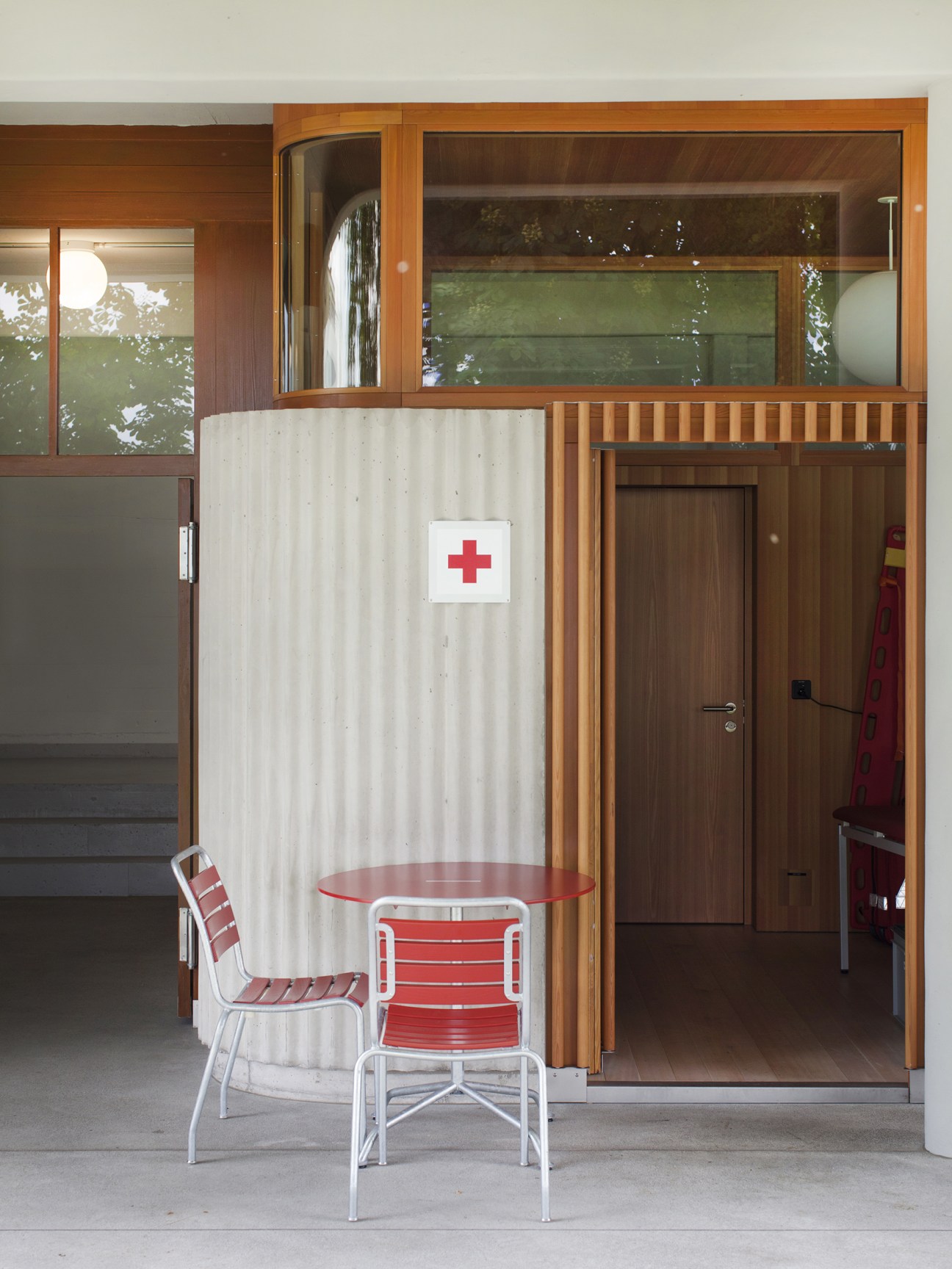 First-aid station at Strandbad Wollishofen with red cross sign, larch screening, and mid-century furniture.