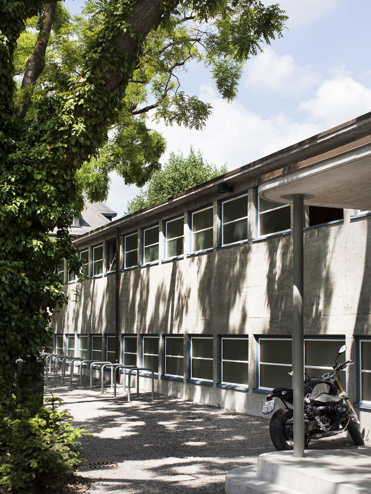 Street-facing side of Strandbad Wollishofen's concrete building with ivy-covered trees, modernist windows and covered parking area.