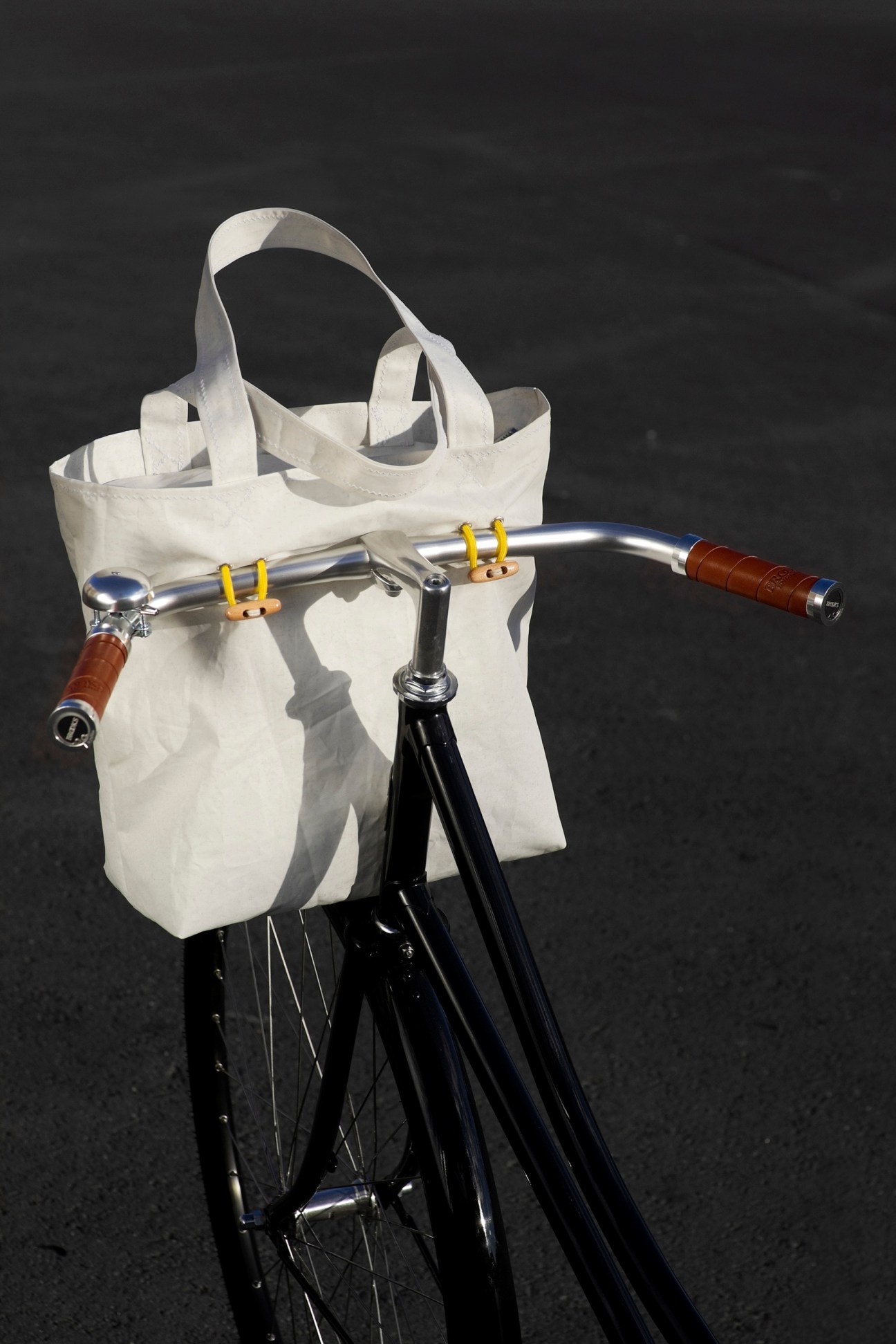 White sail material bike basket with leather handles attached to a bicycle with chrome handlebars.