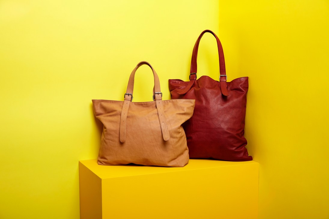 Two leather tote bags in tan and burgundy displayed on a yellow platform against a yellow background.