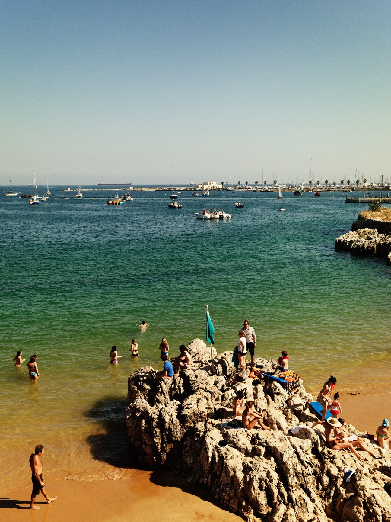 Beachgoers swimming and relaxing on rocks at Queen's Beach in Cascais with sailboats anchored in the bay.