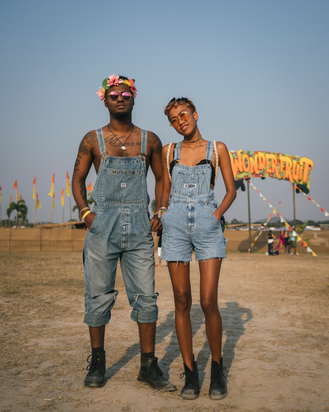 Two festival-goers wearing matching denim dungarees and flower crowns stand at Wonderfruit festival in Thailand.