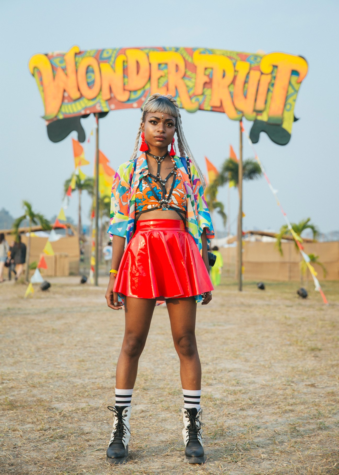 Festival attendee poses in colorful patterned jacket and red skirt beneath Wonderfruit festival sign.