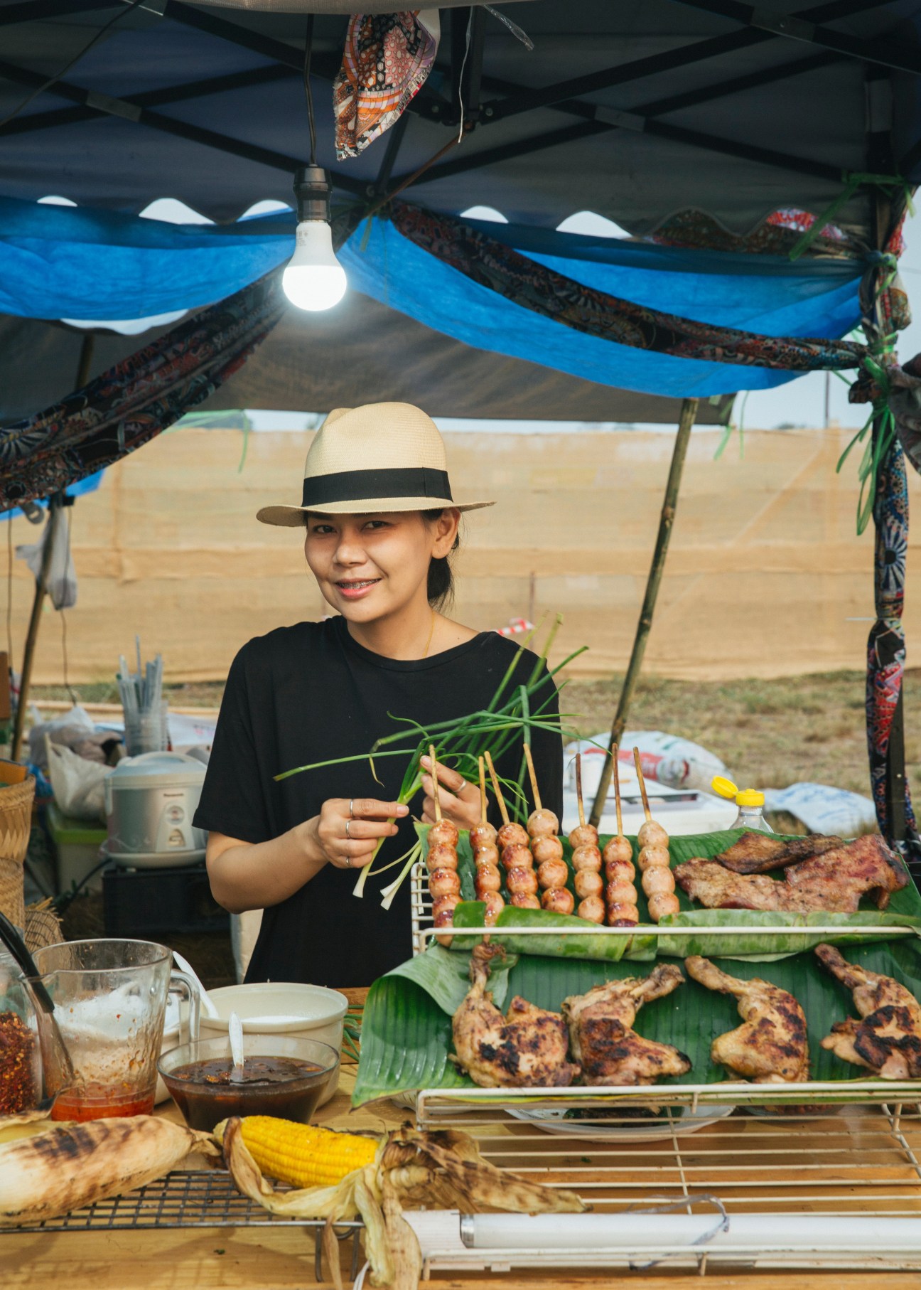 Woman in black shirt and Panama hat serving grilled meat skewers under a blue fabric canopy at Wonderfruit festival.